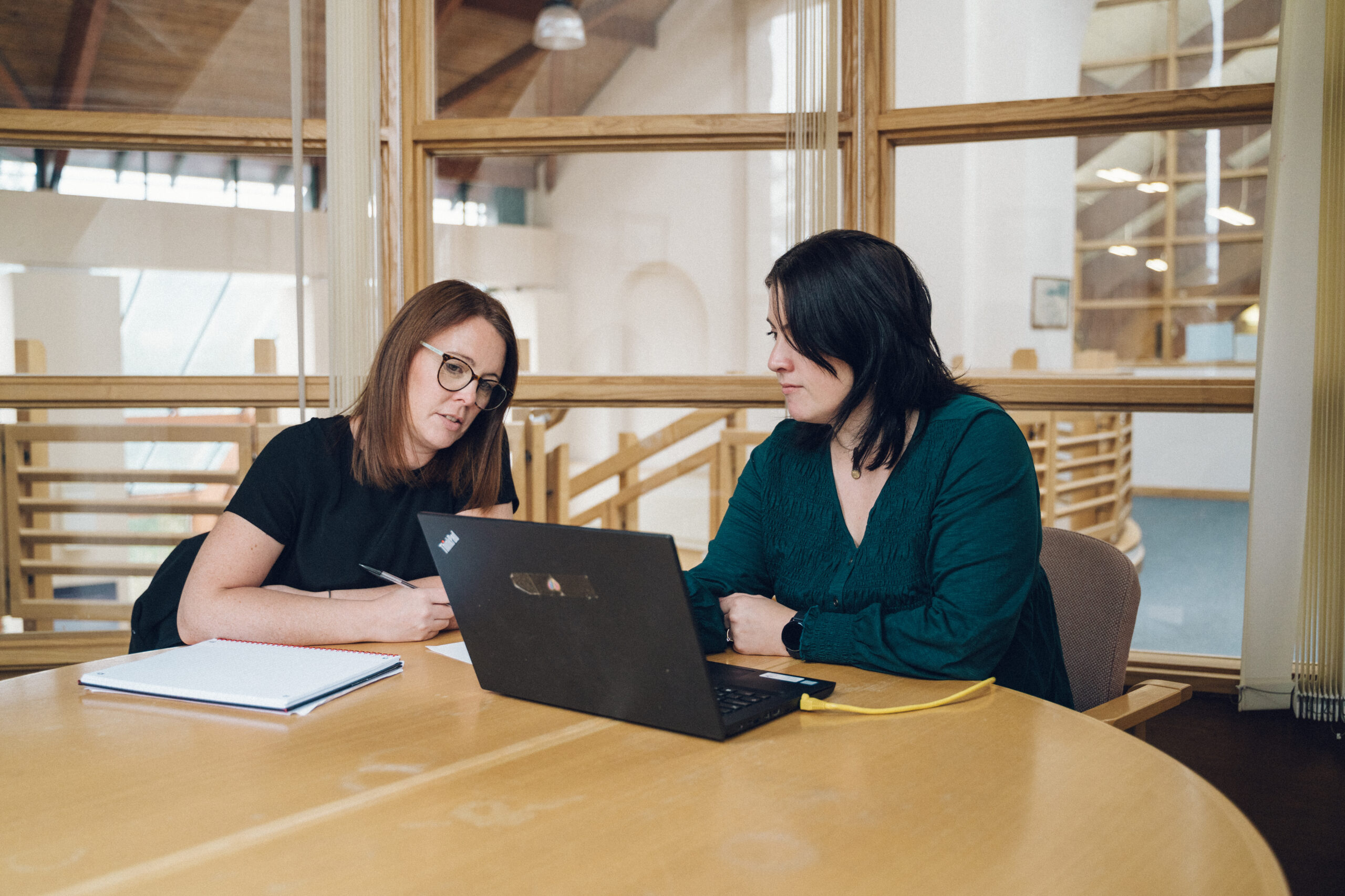 Two women seated at a table in a modern office, collaborating over a laptop with notebooks beside them.