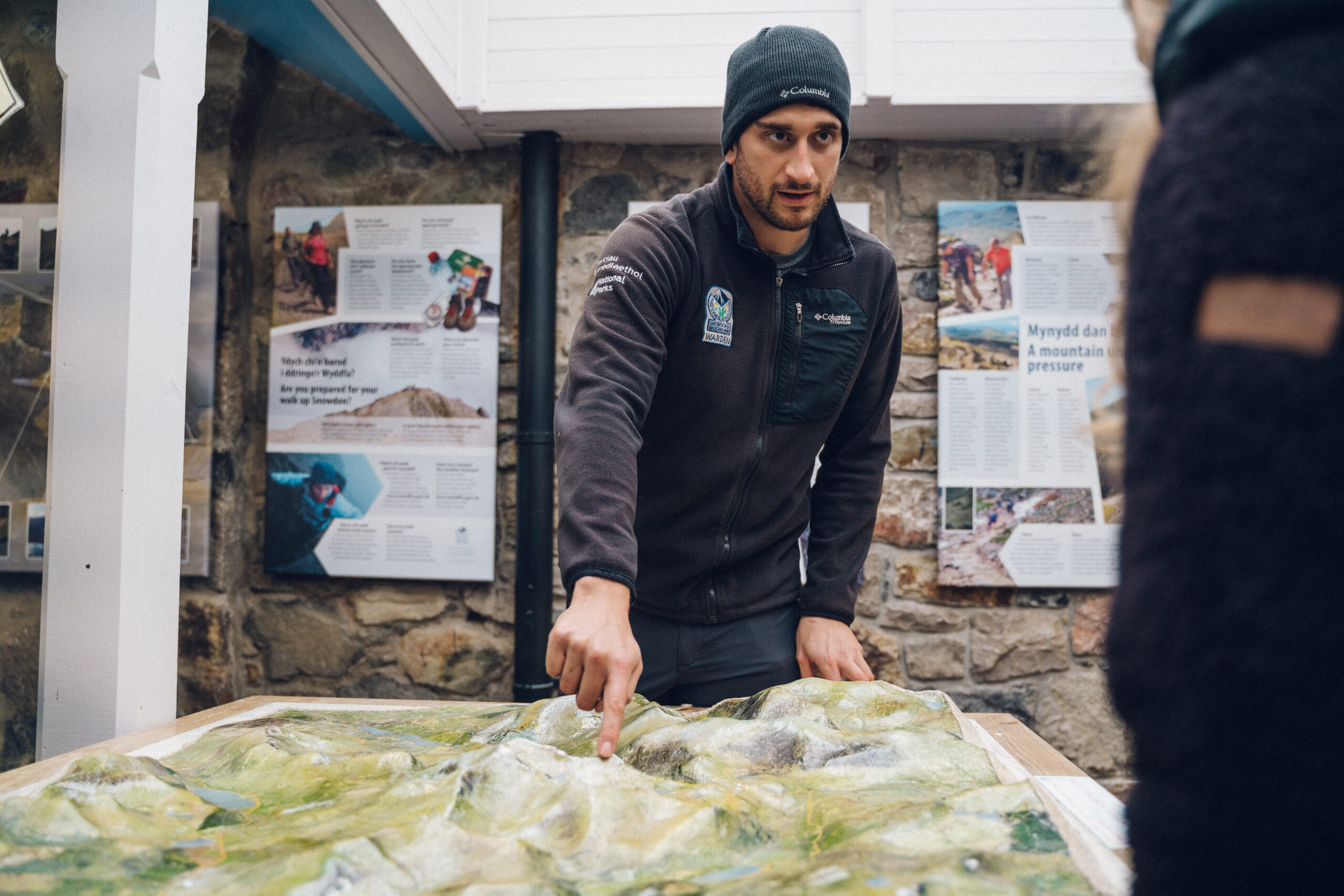 
Warden y Parc Cenedlaethol yn gwisgo het wlanog a siaced awyr agored, yn pwyntio at fap dan do, gyda phaneli gwybodaeth a wal garreg yn y cefndir.  A National Park Warden in a beanie and outdoor jacket is pointing at a raised map display indoors, with informational panels and a stone wall in the background.